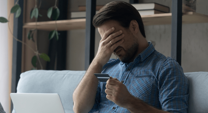 Upset desperate man having problems with wasting money, overspending,, scam and digital fraud. Frustrated disappointed shopaholic, bankrupt holding credit card at laptop, touching face