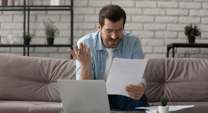 What an outrage. Annoyed young man sit on sofa work with papers at home office read mail letter with bad news unexpected debt. Anxious self employed guy worried by bank report having financial problem